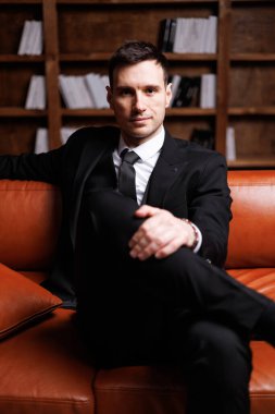 Elegant young man in black suit posing in a modern studio interior with bookshelves and leather sofa, stylish business portrait