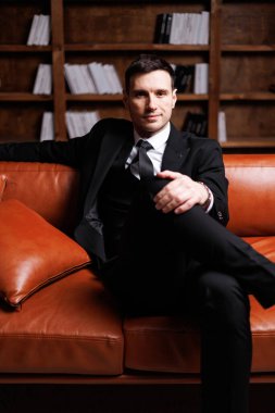 Elegant young man in black suit posing in a modern studio interior with bookshelves and leather sofa, stylish business portrait