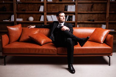Elegant young man in black suit posing in a modern studio interior with bookshelves and leather sofa, stylish business portrait