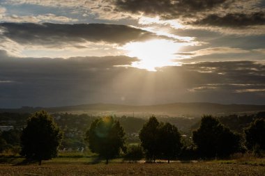 Dramatic sunset with sun rays breaking through thick clouds over a rural valley, illuminating the landscape with trees in the foreground and a town in the distance.