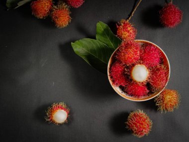 Bir yığın Rambutan meyvesi siyah arka planda beyaz bir kasede ve tahtada birkaç yeşil yaprak var. Tropikal meyve. Rambutan 'ın tatlı meyveleri