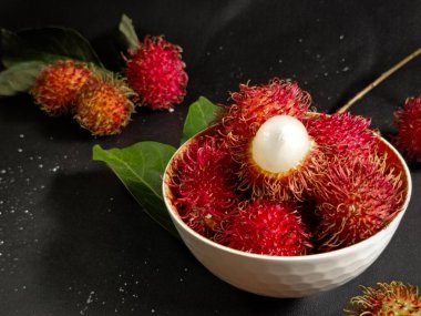 Bir yığın Rambutan meyvesi siyah arka planda beyaz bir kasede ve tahtada birkaç yeşil yaprak var. Tropikal meyve. Rambutan 'ın tatlı meyveleri