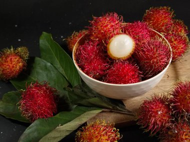 Bir yığın Rambutan meyvesi siyah arka planda beyaz bir kasede ve tahtada birkaç yeşil yaprak var. Tropikal meyve. Rambutan 'ın tatlı meyveleri