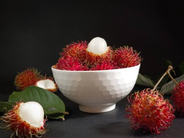 Bir yığın Rambutan meyvesi siyah arka planda beyaz bir kasede ve tahtada birkaç yeşil yaprak var. Tropikal meyve. Rambutan 'ın tatlı meyveleri