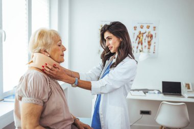 Kadın doktor, yetişkin yaralı bir kadına boyun ortopedik tasması takıyor. Kıdemli bir kadının bayan doktorun muayenehanesini ziyaret ederken fotoğrafı. Doktor, yaşlı bir kadına boyun yaralanmasında yardım ediyor.