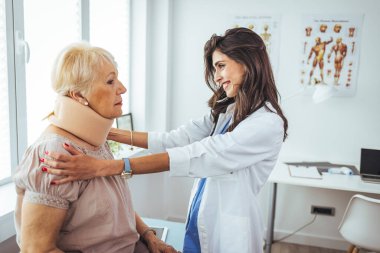 Kadın doktor, yetişkin yaralı bir kadına boyun ortopedik tasması takıyor. Boyun yaralanması yüzünden doktordaki kadın acı çekiyor. Doktor, boyun tasması olan yaşlı bir hastaya reçete yazıyor.