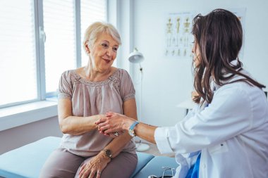 Doktor umut veriyor. Genç kadın hekimin avuçlarında elini tutan yaşlı bayanın gülümsemesini yakından çek. Beyaz önlüklü kadın bakıcı yaşlı insanı cesaretlendiriyor.
