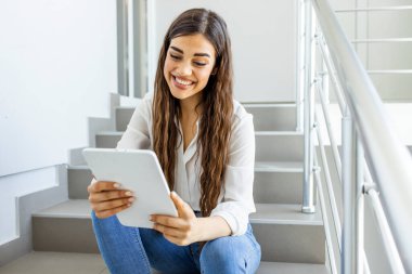Merdivenlerde elinde tabletle oturan bir kız öğrenci. Merdivende tablet kullanan genç bir üniversite öğrencisi. Tablet kullanan üniversiteli kız. Güncel olaylar ile güncellenmeye devam ediliyor