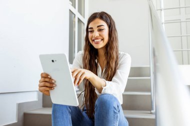 Dijital tablet kullanarak çalışan çekici genç bir kadın ve ofis arka planında otururken gülümsüyor. Kadın merdivenlerde tablet cihazıyla oturuyor. Serbest Çalışan