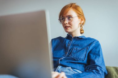 Dizüstü bilgisayarı ve gözlüğü olan genç bir kadın evdeki koltukta oturuyor. 
