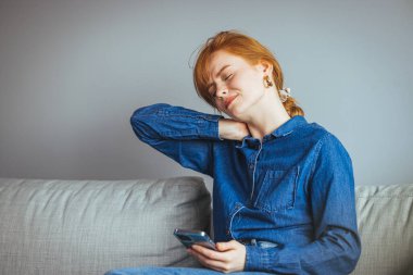 Hüsrana uğramış, boynu ağrıyan genç bir kadın kanepede otururken boynuna masaj yapıyor.