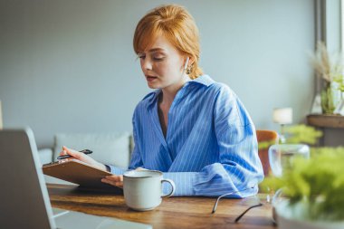 Doymuş ev hanımı, gözlüklü, dizüstü bilgisayarının yanında oturmuş, elinde kalem tutarak aile bütçesi planlaması yapıyor.