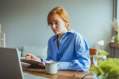 Doymuş ev hanımı, gözlüklü, dizüstü bilgisayarının yanında oturmuş, elinde kalem tutarak aile bütçesi planlaması yapıyor.