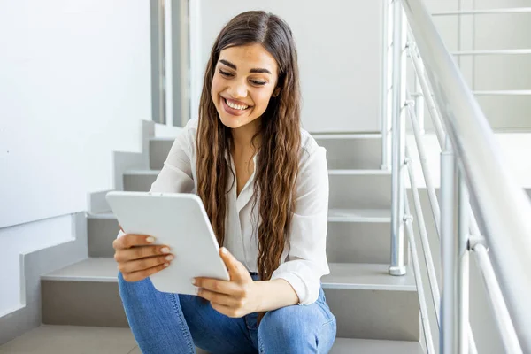 Female student sitting on stairs with a tablet pc. Young female college student using tablet on a staircase. College girl using tablet. Staying updated with current affairs