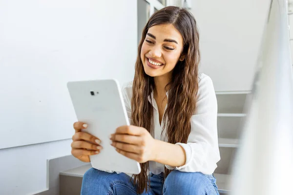 Female student sitting on stairs with a tablet pc. Young female college student using tablet on a staircase. College girl using tablet. Staying updated with current affairs