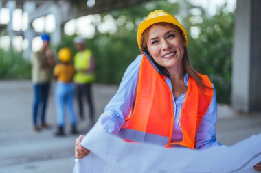 Beyaz kadın işçi akıllı telefon kullanıyor ve inşaat alanının planlarını tutuyor.