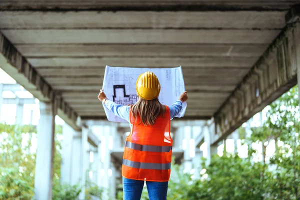 Back view of female construction worker in yellow safety helmet holding blueprint at the construction site