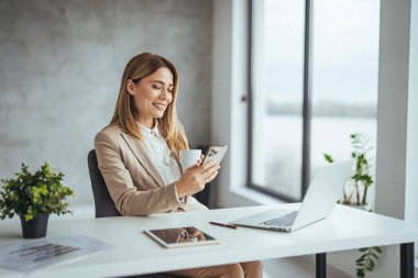 Ofisteki dizüstü bilgisayarda çalışırken akıllı telefon kullanan ve çay ya da kahve içen Kafkasyalı genç iş kadını.