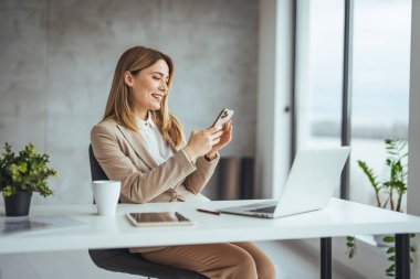 Ofisteki dizüstü bilgisayarda çalışırken akıllı telefon kullanan ve çay ya da kahve içen Kafkasyalı genç iş kadını.