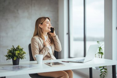 Kafkasyalı genç iş kadını akıllı telefonla konuşuyor ve ofisteki bilgisayarında çalışıyor.