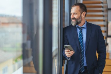 Yakışıklı iş adamı gözlük takıyor akıllı telefon kullanıyor ve ofis penceresinin yanında dururken gülümsüyor, 