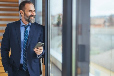 Modern bir ofiste akıllı telefon kullanan genç bir iş adamının fotoğrafı. Olgun bir tasarımcının ofisteki bir cep telefonuyla mesajlaşırken görüntüsü. Başlangıç ihtiyaçlarını karşılamak için en akıllı teknolojiyi kullanıyor.