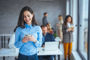 Güzel iş kadını toplantıyı beklerken mesaj atıyor. Genç bir iş kadınının iş yerinde telefonunu kullanması. Ofiste sıradan giyinmiş gülümseyen kadın. Elinde cep telefonu olan bir iş kadını 
