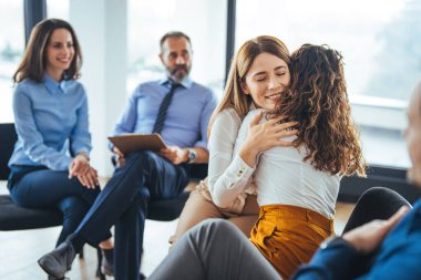 Destek grubu toplantısı için toplanıyor. İki kişi birbirini kucaklıyor ve diğer üyeler de onları destekliyor. Çok ırklı bir yetişkin grubu grup terapisine katılıyor..