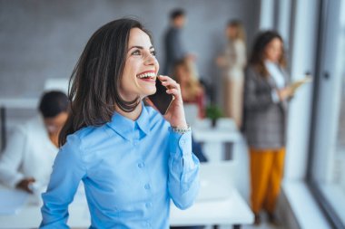 Akıllı telefonu tutarken kameraya gülümseyen genç bir iş kadını. Arka planda meslektaşları olan neşeli genç bir iş kadını. Sadece bir arama önemli bir şey getirdi.