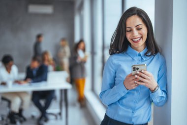 Ofiste genç bir iş kadını ve akıllı telefondan mesaj atıyor. Modern ofiste cep telefonu kullanan güzel bir iş kadını. Akıllı telefonlu bir iş kadını pencerenin yanında oturuyor ve ofiste boş zamanlarında eğleniyor..