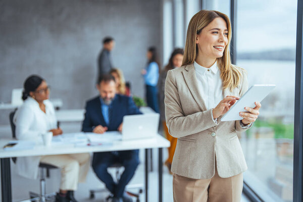 Городская счастливая бизнес-леди пользуется планшетным компьютером и работает. Счастливая деловая женщина с цифровым планшетом. Молодая ведущая бизнесвумен, использующая беспроводной планшет. Дизайнер, стоящий в ее офисе с помощью онлайн-приложения