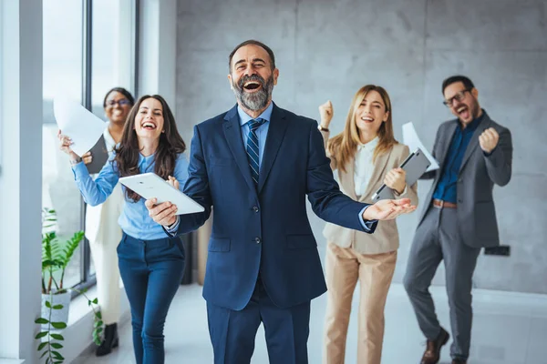 Successful business group celebrating an achievement at the office with arms up and looking at the camera smiling. Shot of a group of young businesspeople huddled together in solidarity in a modern office