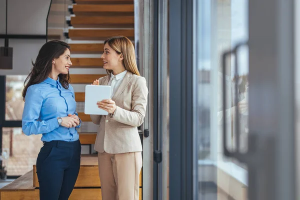 Two businesswomen chatting, meet together in hallway, share opinions and professional information, discuss project having business conversation or informal talk with tablet and coffee mug in office