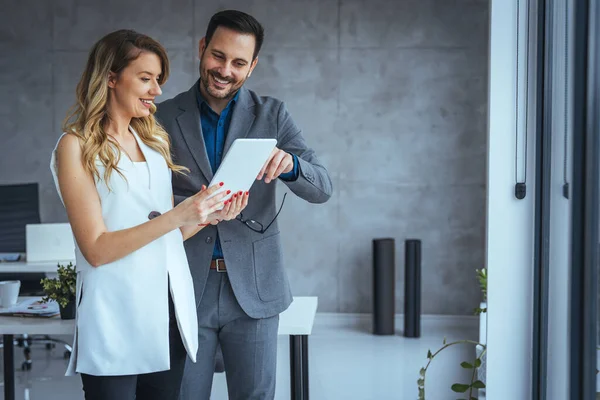 Two happy colleagues smiling cheerfully in an office. Happy mature businessman holding a smartphone while standing with his female colleague in a modern workplace. Businesspeople working as a team.