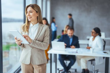 Dijital tabletle ofiste duran genç bir iş kadınının portresi. Ofiste kendine güvenen bir iş kadını. Modern ofiste dijital tablet bilgisayarda çalışan güzel bir iş kadını.