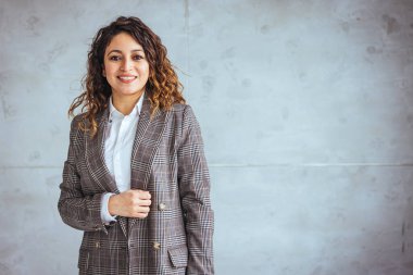 Modern bir ofiste duran kendine güvenen genç bir iş kadınının fotoğrafı. Gri arka planda stüdyonun vesikalığı. İspanyol modeli. Çapraz kollarla gülümseyen güzel kadın 