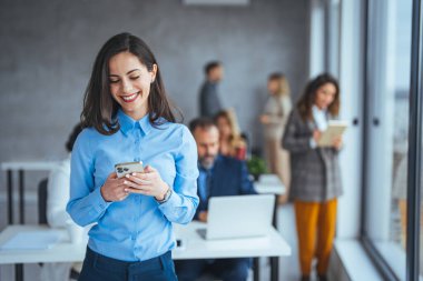 Ofiste genç bir iş kadını ve akıllı telefondan mesaj atıyor. Modern ofiste cep telefonu kullanan güzel bir iş kadını. Akıllı telefonlu bir iş kadını pencerenin yanında oturuyor ve ofiste boş zamanlarında eğleniyor..