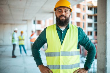 Arka planda bulanık iş arkadaşlarıyla birlikte inşaat alanında çalışan elleri kalçalı beyaz mühendisin portresi.