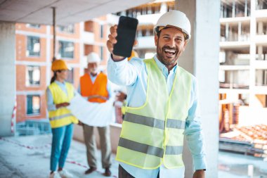 Planları elinde tutan ve akıllı telefon gösteren bir mühendis. Erkek inşaat mühendisi akıllı telefonunu kameraya gülümseyerek gösteriyor. Kasklı erkek mühendis akıllı telefonu gösteren kameraya bakıyor.