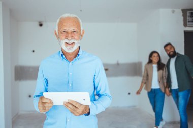 Yeni evinde elinde dosyaları tutan gülümseyen emlakçının portresi. Arka planda mutlu çift dururken kameraya bakan üst düzey bir emlakçının portresi..