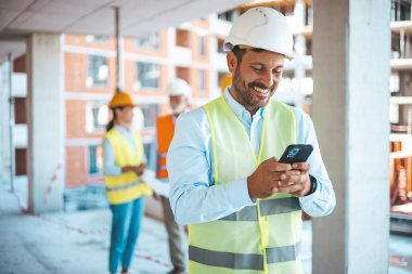 İnşaat alanında akıllı telefonunu kullanan genç bir işadamını vurdu. İnşaat alanında duran beyaz kasklı genç mühendis portresi. İşçi, internet ve sosyal medya için gülümsüyor ve akıllı telefon tutuyor.. 