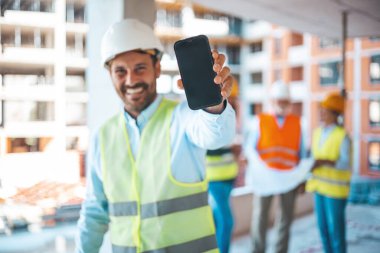 Planları elinde tutan ve akıllı telefon gösteren bir mühendis. Erkek inşaat mühendisi akıllı telefonunu kameraya gülümseyerek gösteriyor. Kasklı erkek mühendis akıllı telefonu gösteren kameraya bakıyor.