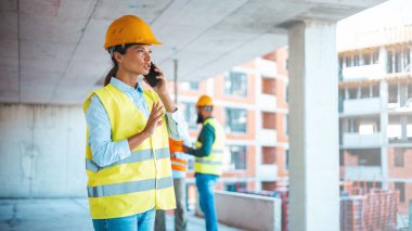 Sert şapkalı kadın mühendis cep telefonu kullanıyor, endüstriyel işçi cep telefonu kullanıyor. İnşaat alanında çalışırken akıllı telefon kullanan genç bir kadının fotoğrafı.