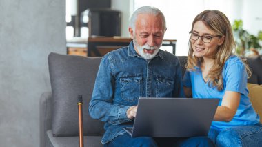 Kadın hemşire, gün boyunca kanepede otururken üst düzey erkek hastasına danışıyor. Aktif kıdemli bir adam dizüstü bilgisayar kullanıyor ve kadın bakıcı evdeki kanepede oturuyor. Hemşire dizüstü bilgisayarla çalışan yaşlı emeklilere yardım ediyor