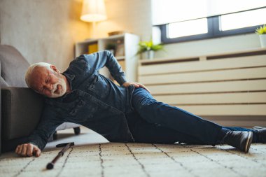 Yaşlı kıdemli adam kayıyor ve düşüyor. Oturma odasında düşen yaşlı insan. Hüzünlü, modaya uygun, kareli tişört giyen yaşlı adam beyaz ışıklı modern iç mekanda bastona ve divana yaslanarak kalkmaya çalışıyor.