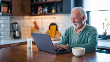 Evde dizüstü bilgisayarda çalışan kıdemli bir adam. Emekli adam mutfaktaki evinde iyi haberi kutlamak için dizüstü bilgisayar kullanıyor. Ev mutfağında çay fincanı ve dizüstü bilgisayarla kıdemli bir yetişkin..
