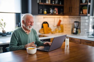 Modern mutfakta bilgisayarı olan bir adam. Evdeki dizüstü bilgisayarla internete bağlanan son sınıf öğrencisi. Emeklilik üzerine uzaktan çalışma. Gülümseyen kıdemli adam evdeki mutfakta dizüstü bilgisayar kullanıyor.