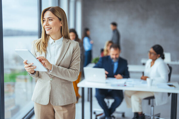 Портрет молодой предпринимательницы, стоящей в офисе с цифровым планшетом. Уверенная деловая женщина в офисе. Красивая деловая женщина стоит и работает на цифровом планшетном компьютере в современном офисе
