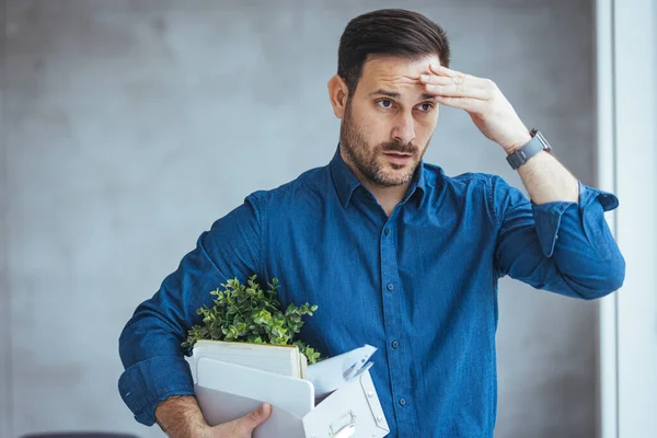Business, Firing and Job Loss Concept - Sad Fired Male Office Worker With Box of His Personal Stuff. Fired male employee holding box of belongings in an office. Displeased mid adult businessman leaving the office
