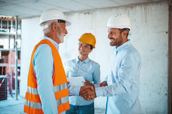 Woman architect or engineer looking at male colleagues handshaking at construction site 